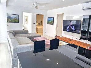 A modern living room with a grey sectional sofa, a TV, and a dining area with a table and chairs. - B8 - 2 Bedrooms Serviced Apartment with pool view, Karon Beach (Tambon Karon)