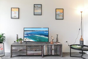 A living room with a flat-screen TV on a wooden cabinet, framed travel posters on the wall, and a floor lamp. - Manly beach: Everything minutes away! (Manly)