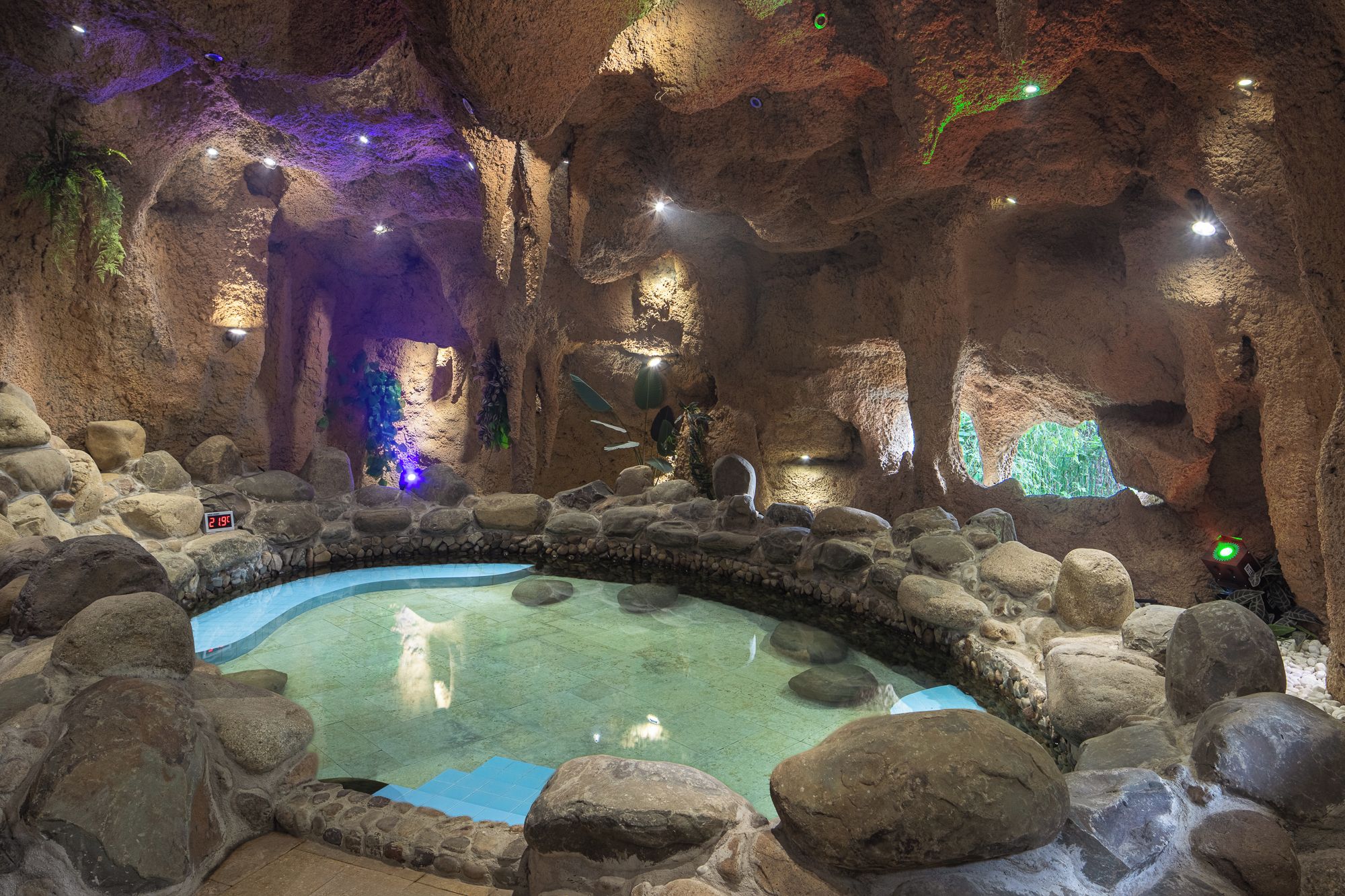 Ein innenliegendes, höhlenähnliches Ambiente mit einem zentralen Pool, der von Felsen umgeben und durch Scheinwerfer beleuchtet wird.