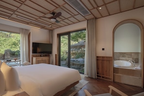 A bedroom with a large bed, a television, a bathroom with a bathtub, and a view of the outdoors.