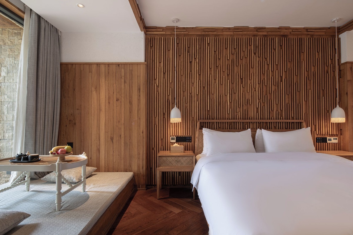 A hotel room with a large bed, wooden panel wall, bedside tables with lamps, a small table with fruits, and a view of a stone wall.