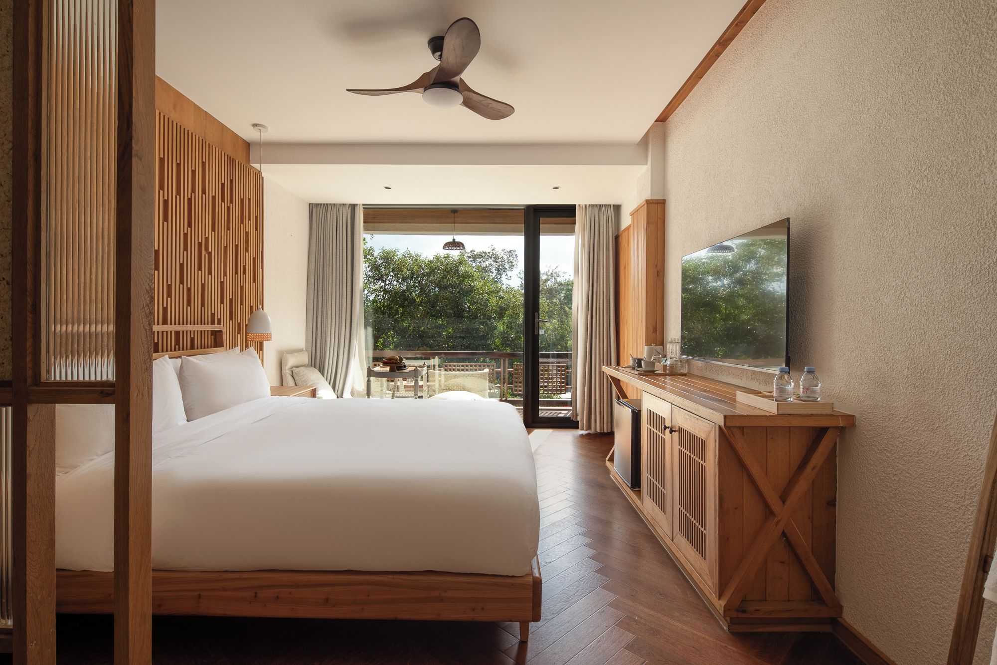 A hotel room with a large bed, a ceiling fan, a TV, and a balcony with a view of greenery.