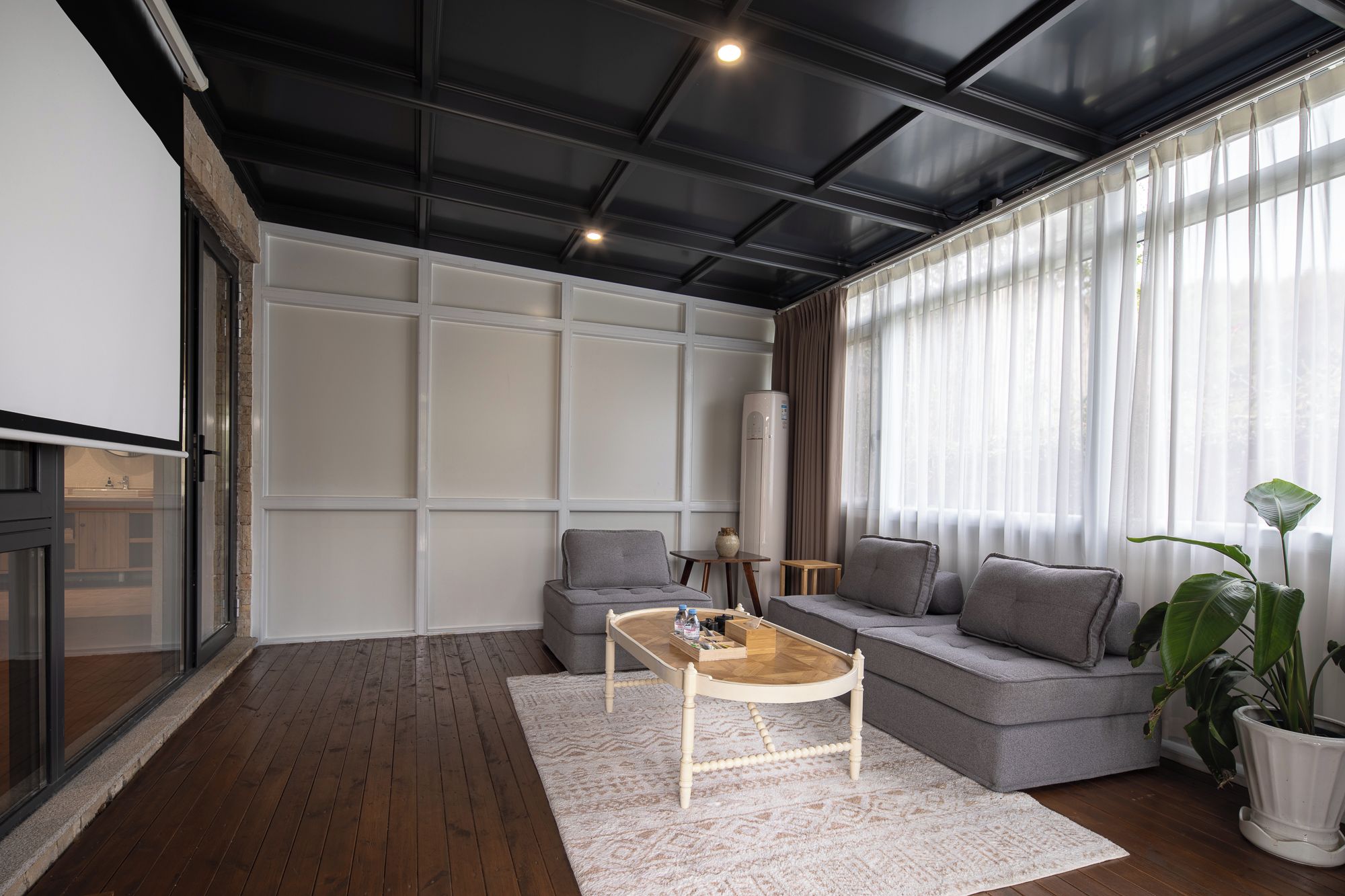 Ein modernes Wohnzimmer mit einem grauen Sofa, einem Holztisch und einem großen Fenster mit transparenten Vorhängen.