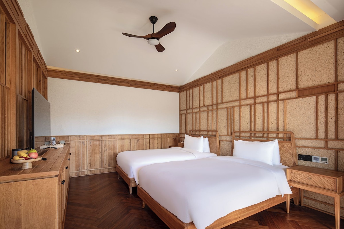 A hotel room with two beds, wooden headboards, a ceiling fan, a flat-screen TV, and a wooden dresser with fruit on it.