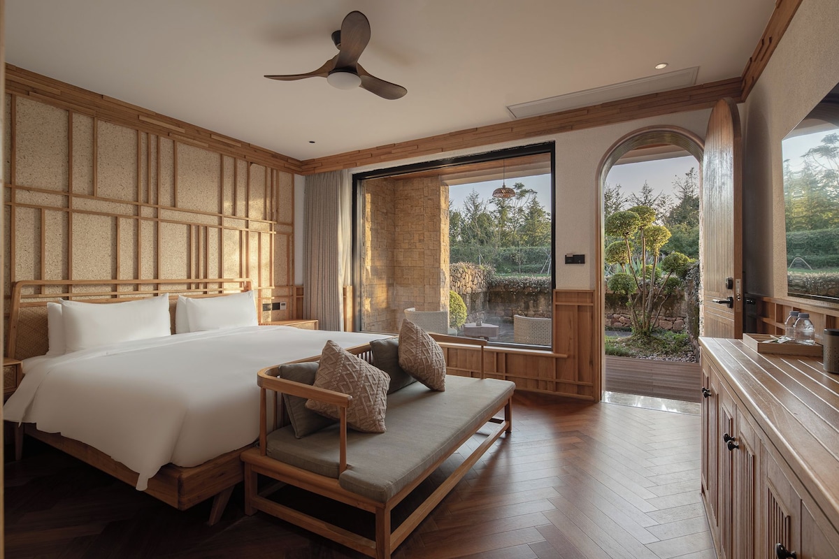 A bedroom with a large bed, a wooden bench with cushions, a ceiling fan, and a view of a garden through a glass door.