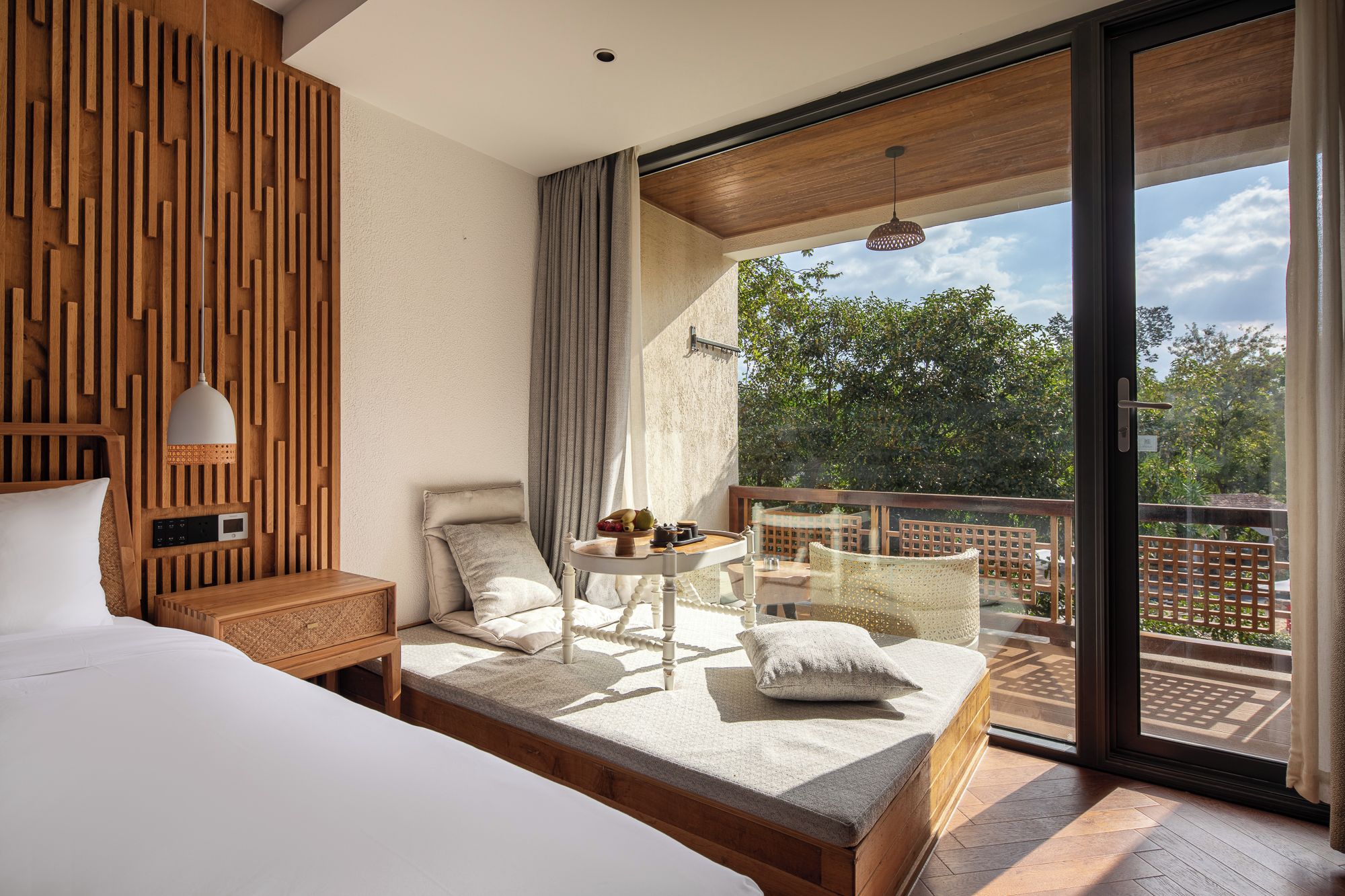 Een moderne slaapkamer met een groot bed, een klein tafeltje met een lamp en een balkon met uitzicht op bomen.