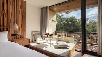Een moderne slaapkamer met een groot bed, een klein tafeltje met een lamp en een balkon met uitzicht op bomen.