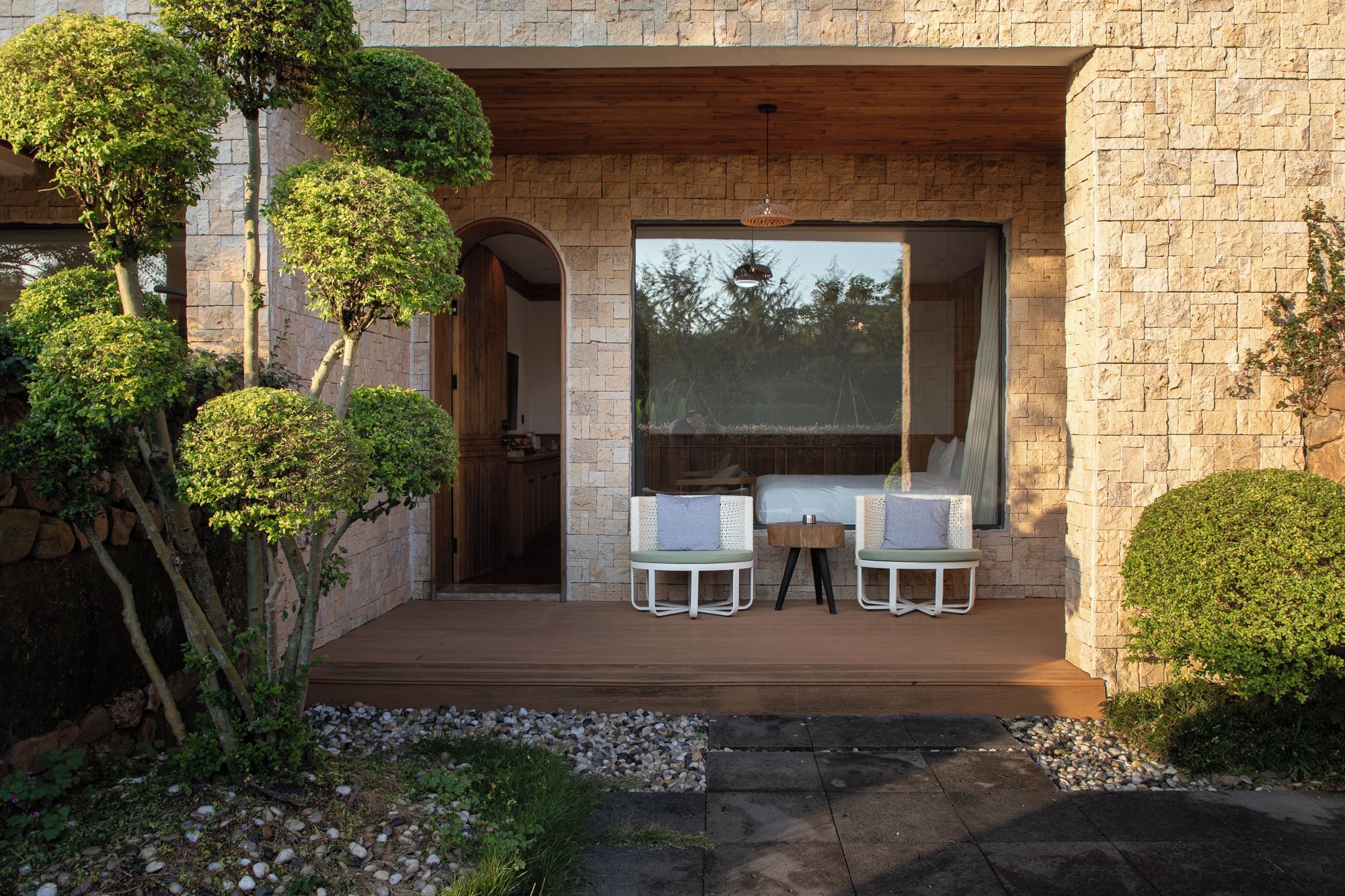 A wooden deck with two chairs and a table, a stone wall, and a large window.