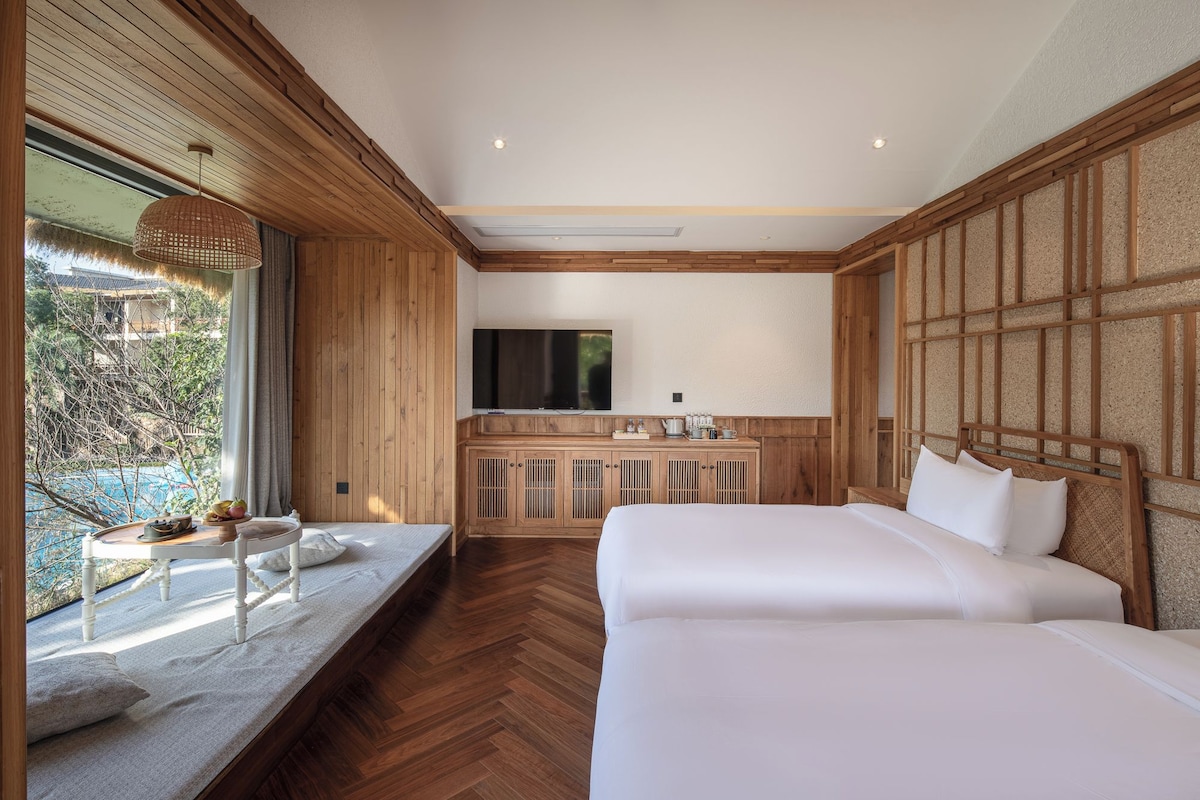 A bedroom with a wooden ceiling, a TV mounted on the wall, a bed, and a window with a view of greenery.