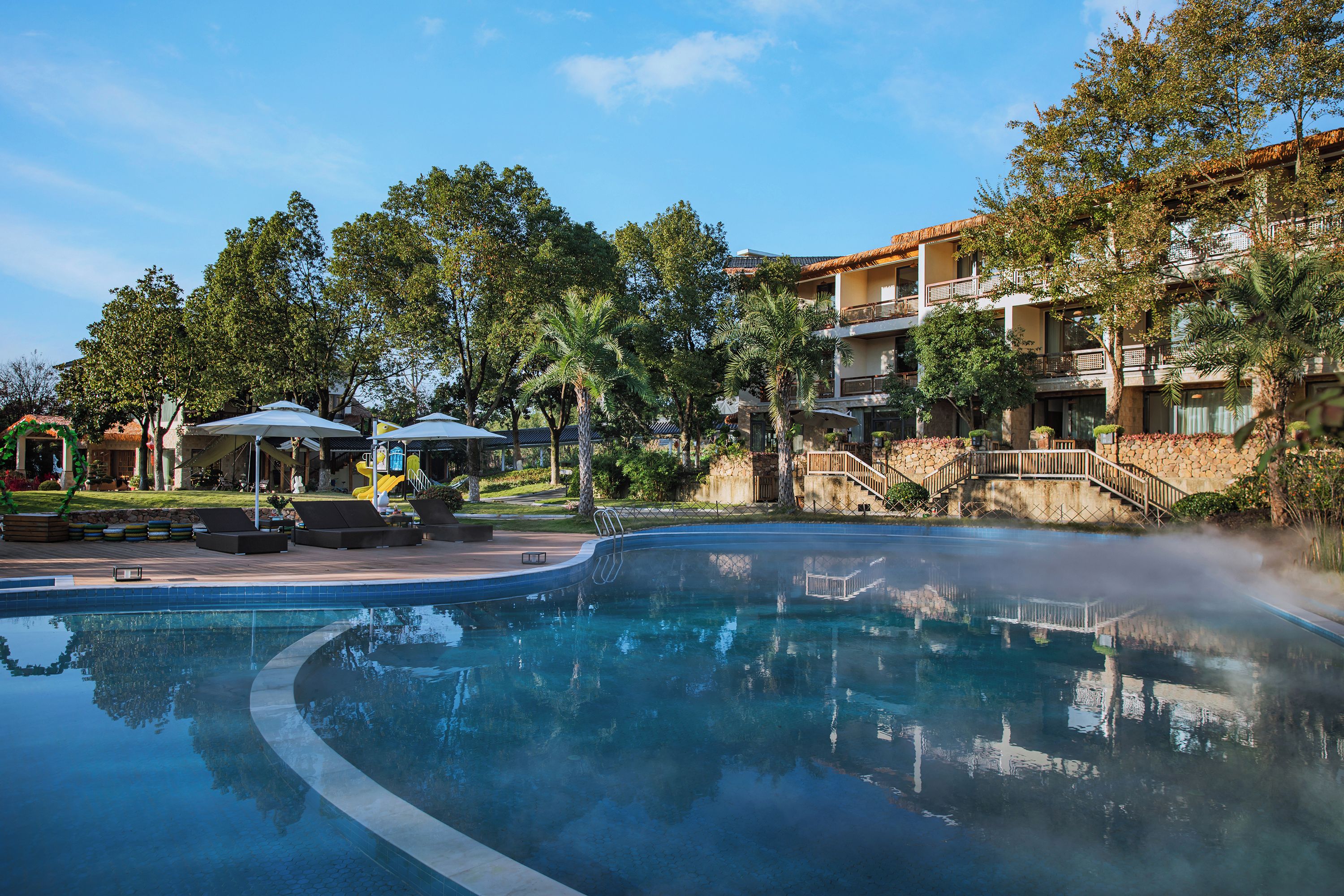 A swimming pool with lounge chairs and umbrellas, surrounded by trees and a building with balconies.