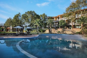 Una piscina amb gandules i ombrel·les, envoltada d'arbres i un edifici amb balcons.