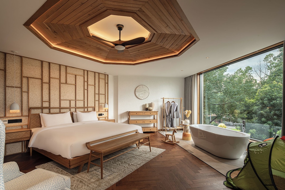 A modern bedroom with a large bed, a wooden ceiling design, a view of greenery outside, and a bathtub.