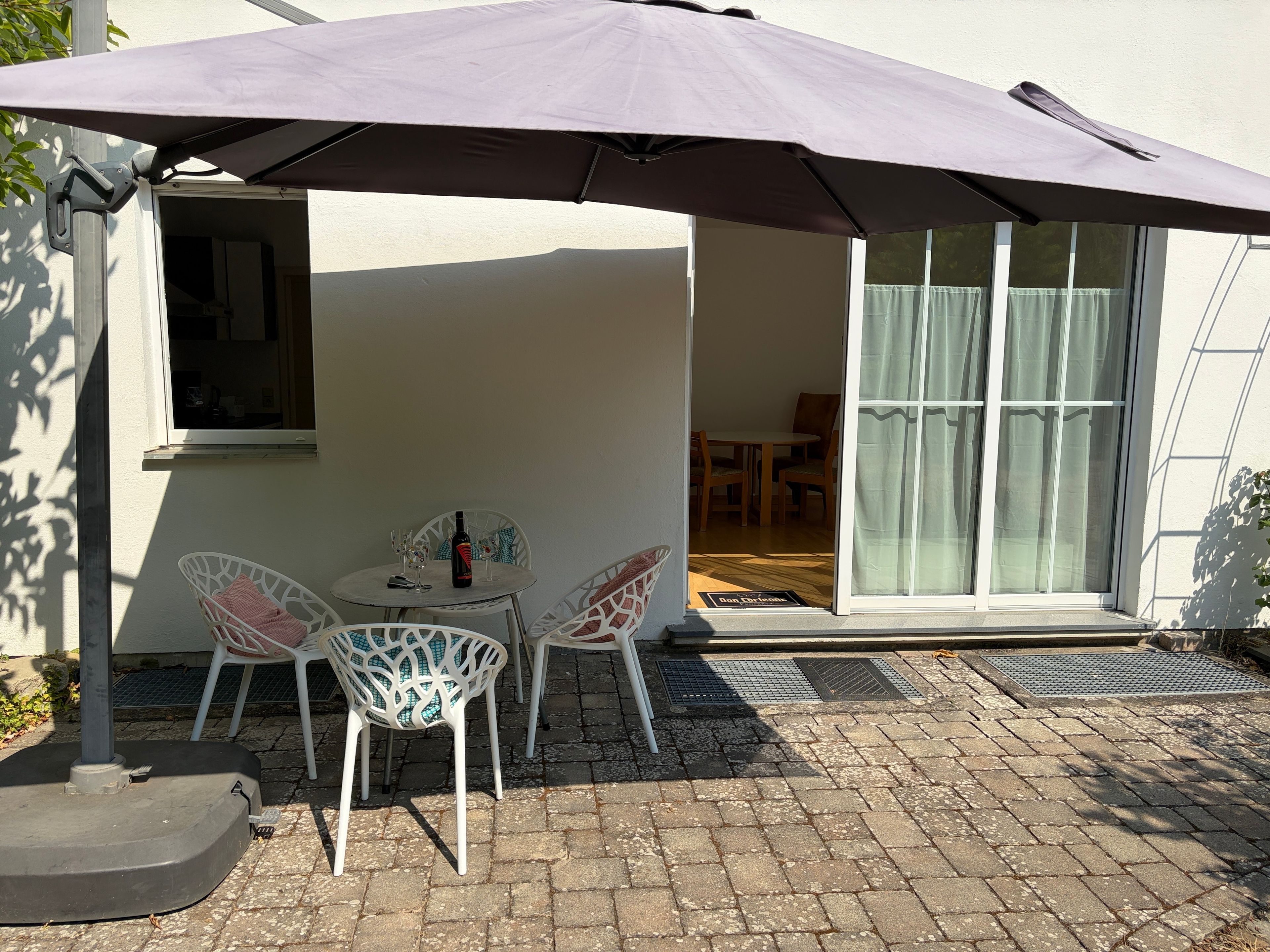 Um pátio externo com mesa e cadeiras sob um grande guarda-sol, porta de correr de vidro que dá para um espaço interno e um pequeno degrau até a porta.