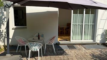 Um pátio externo com mesa e cadeiras sob um grande guarda-sol, porta de correr de vidro que dá para um espaço interno e um pequeno degrau até a porta.