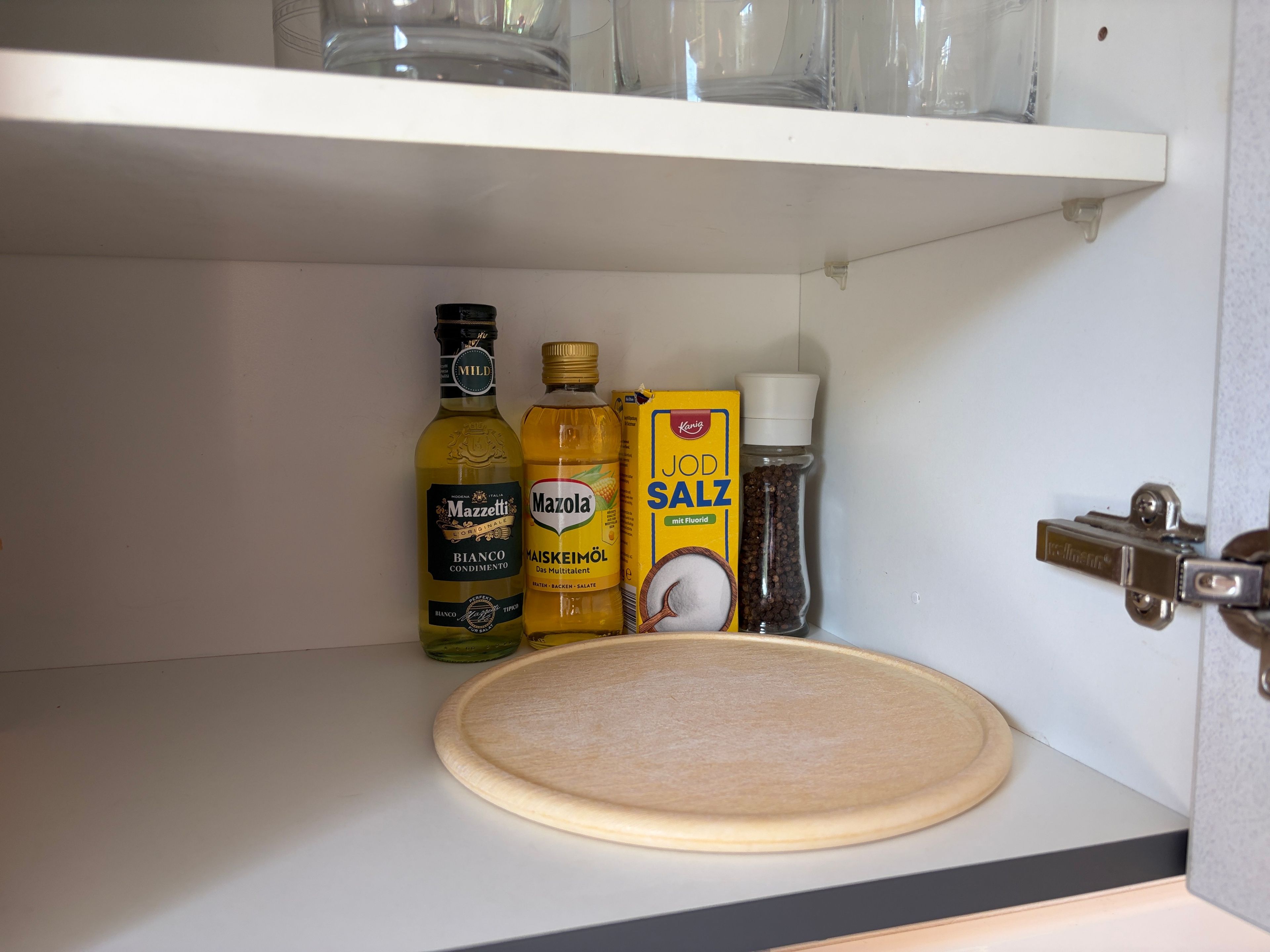 A kitchen cabinet with a wooden cutting board, a bottle of Mazella mayonnaise, a bottle of Mazetti olive oil, a box of Jod Salz salt, and a pepper grinder.