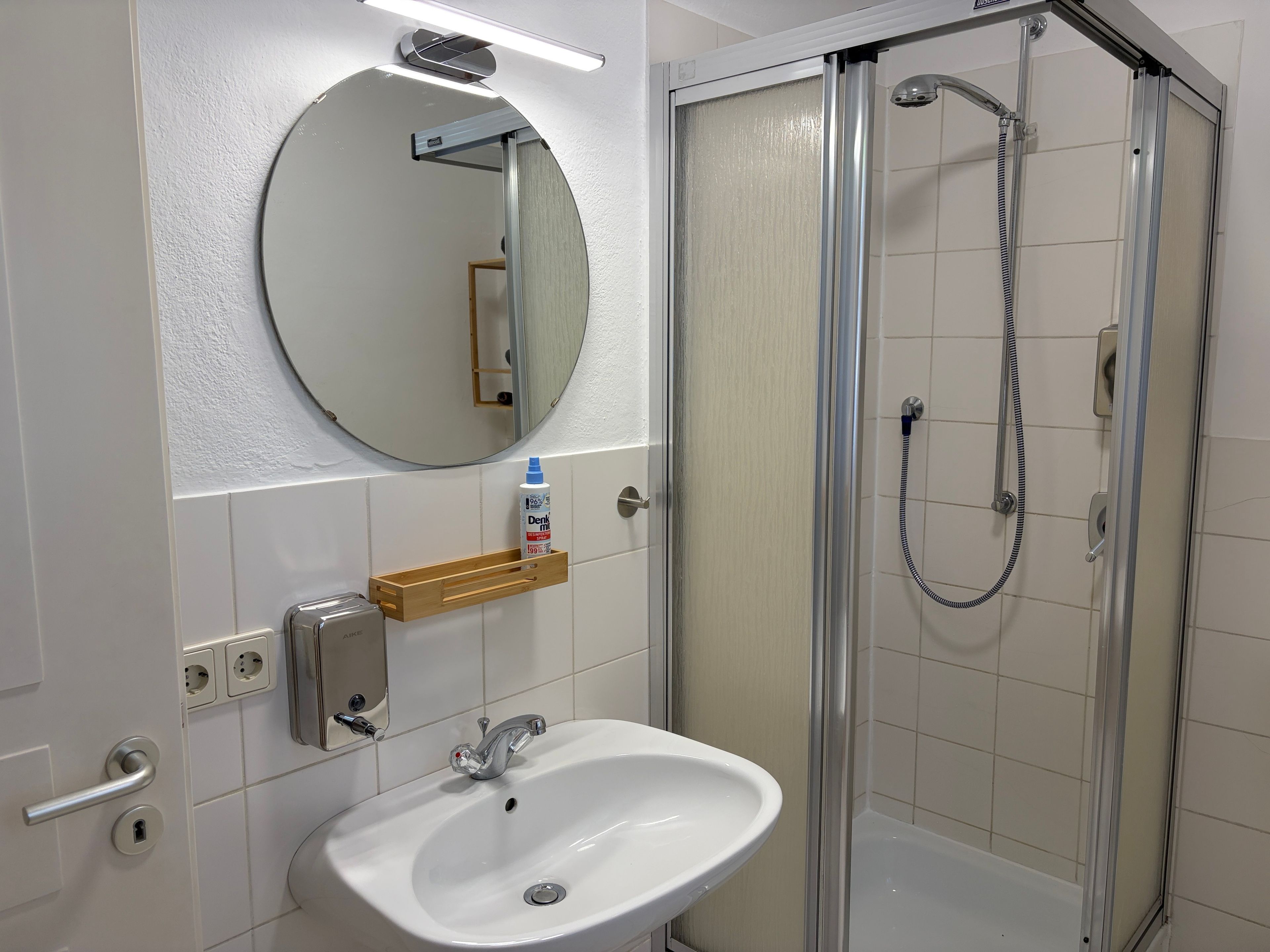 A bathroom with a white sink, a mirror, a shower enclosure, and a wooden shelf with a bottle.