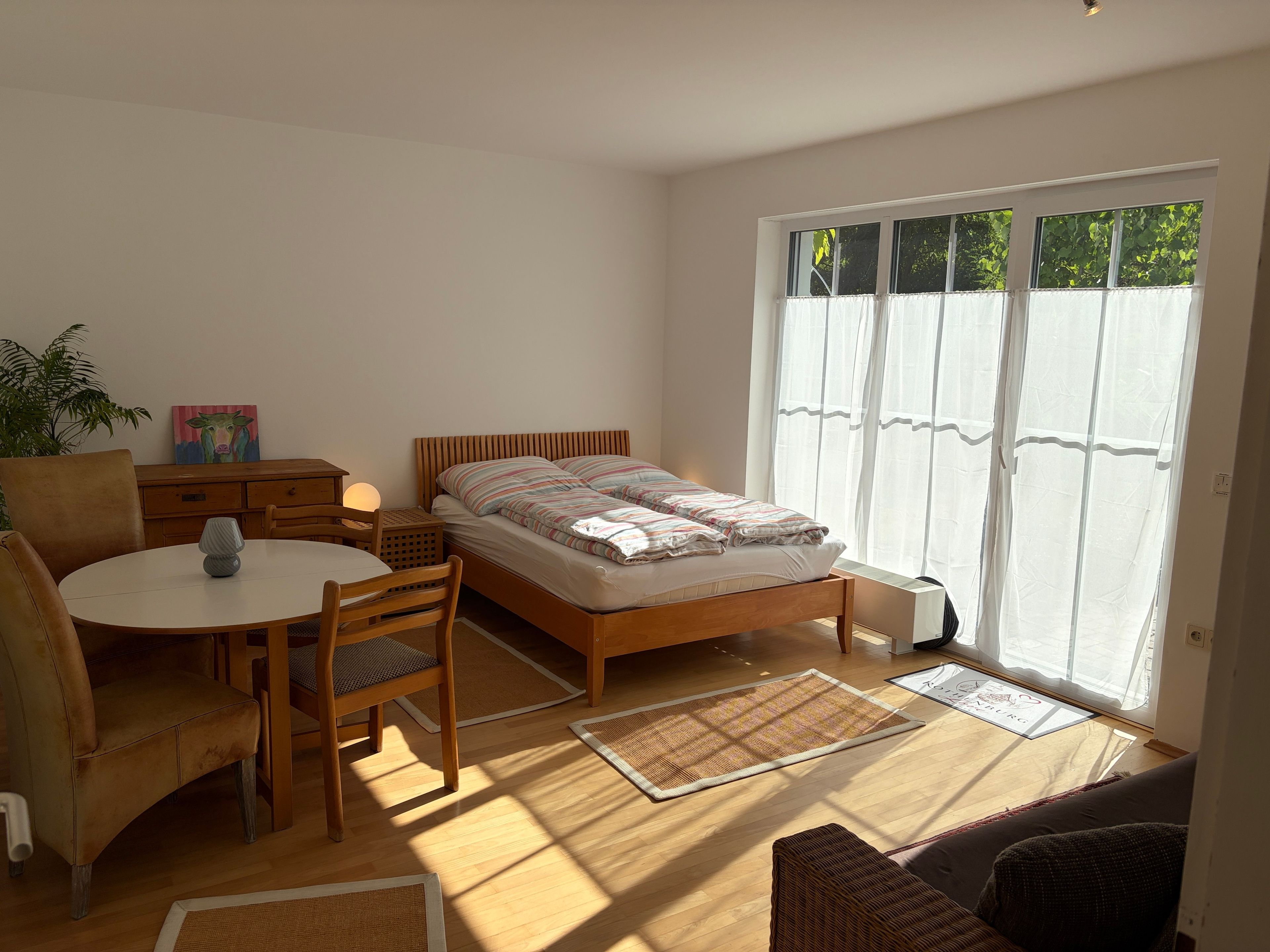 Une chambre à coucher avec un lit, une chaise, une table, une plante et une fenêtre avec des rideaux.