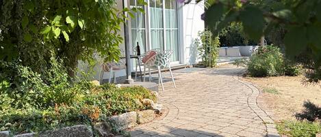 A paved pathway leading to a building with a glass door, surrounded by greenery and a patio area with chairs.