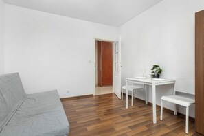 Une pièce au décor minimaliste, avec un canapé gris, une table à manger blanche et des chaises, ainsi qu’une porte en bois.