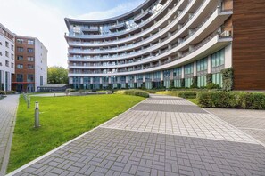 Un bâtiment moderne à plusieurs étages, doté d’une façade incurvée, d’une cour aménagée avec un chemin pavé et d’une zone herbeuse.