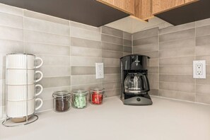 Una cocina con una cafetera, un escurridor para tazas y tres frascos de especias.