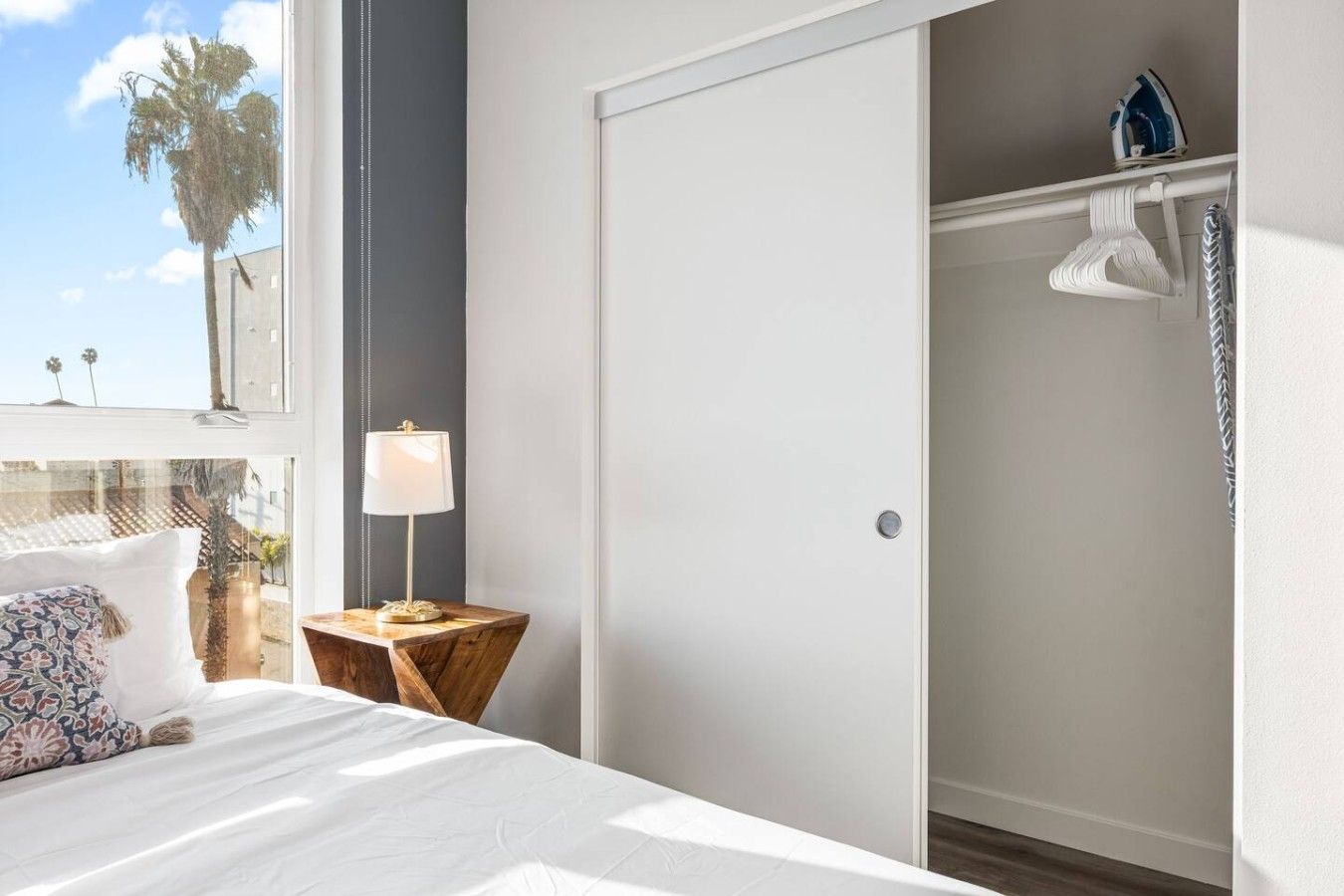 A bedroom with a bed, a nightstand, a lamp, a wardrobe, and a view of palm trees through a window.
