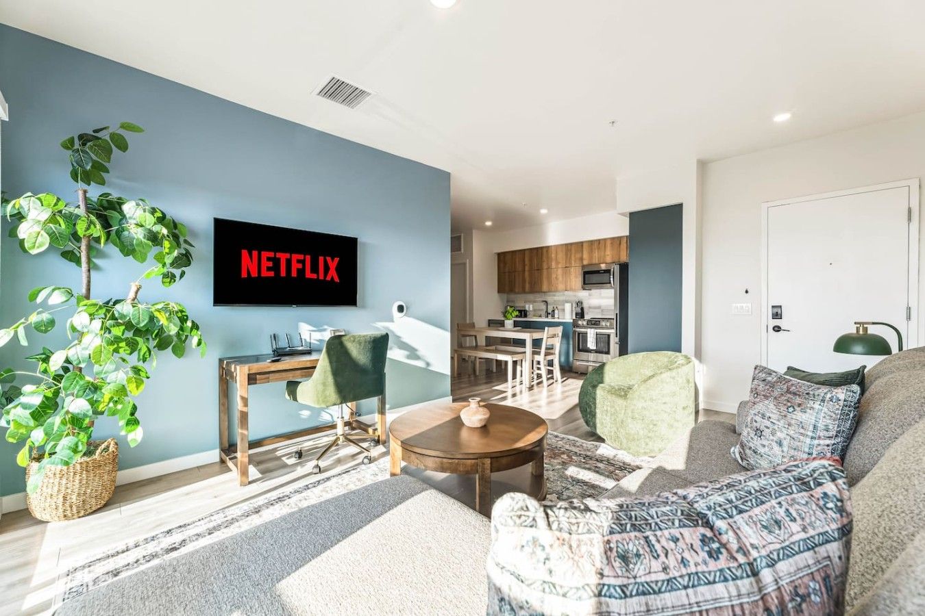 A modern living room with a flat-screen TV, a dining area, and a kitchen in the background.