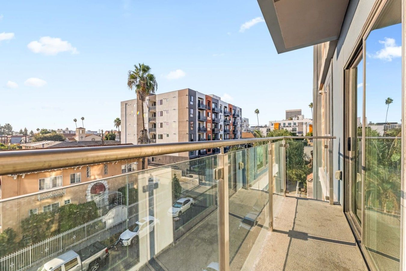 A balcony with a glass railing overlooking a cityscape, including a multi-story building and palm trees.