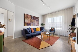 Ein modernes Wohnzimmer mit einer Couch, einem Couchtisch und einem Sessel. An der Wand hängt ein großes abstraktes Gemälde, ein Fenster mit leichtem Vorhang und ein Topf mit einer Pflanze auf der Fensterbank.