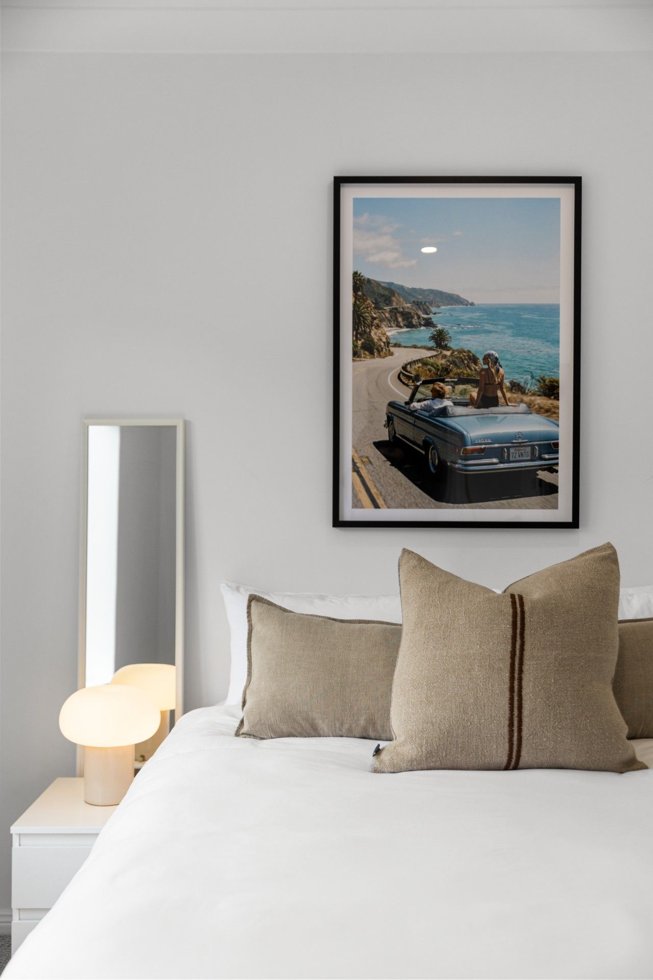 A bedroom with a bed, pillows, a bedside lamp, a framed picture of a car on a coastal road, and a nightstand.