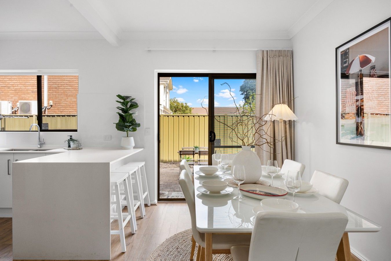 A modern kitchen with a central island, a dining area with a glass table, and a sliding glass door leading to an outdoor patio.