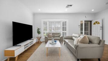 A modern living room with a flat-screen TV, two sofas, a coffee table, and a large window with blinds.