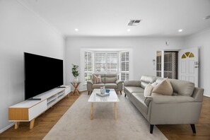 Ein modernes Wohnzimmer mit einem Flachbildfernseher, zwei Sofas, einem Couchtisch und einem großen Fenster mit Jalousien.
