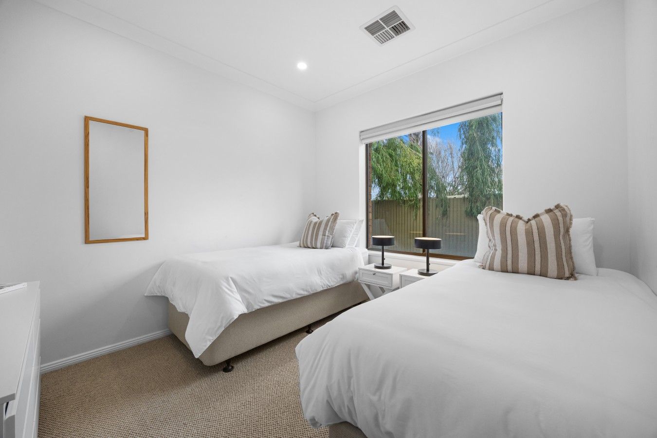 A bedroom with two beds, a window with a view of greenery, and a white ceiling with a vent.