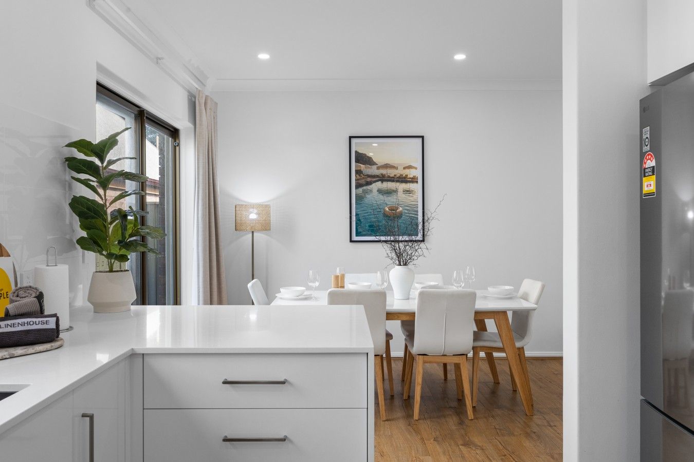 A modern kitchen with white cabinetry, a dining area with a table set for four, and a large window with a view of the outdoors.