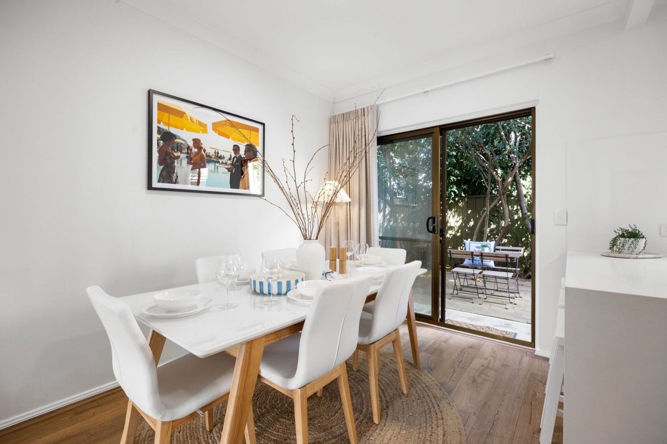 A dining area with a white table set for four, a vase with branches, and a painting above.
