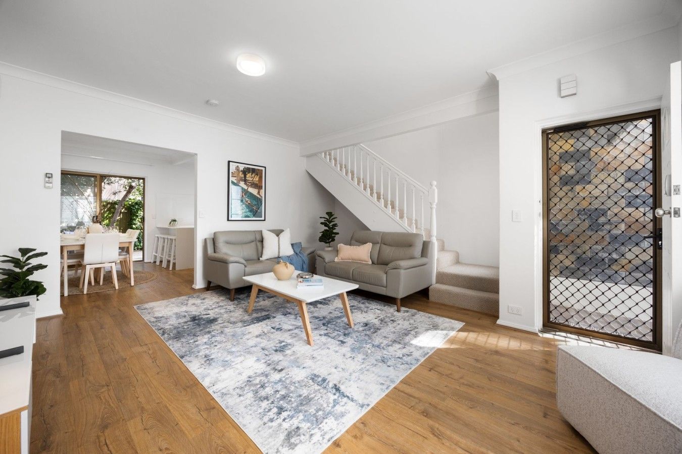 A modern living room with a staircase, a dining area, and a view through a glass door.
