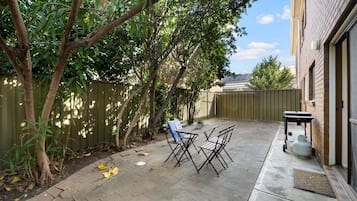 Un jardin avec une terrasse, une table, des chaises, une bouteille de gaz et une clôture.