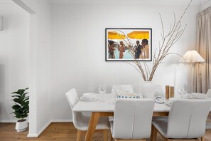 A dining area with a white table set for four, a framed picture above the table, and a vase with branches on the right.