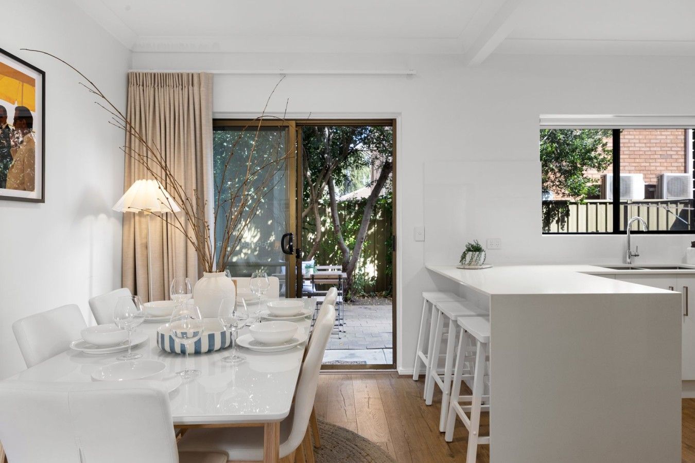 A modern kitchen with white cabinetry, a dining area with a table set for four, and a view of the outdoors through a glass door.