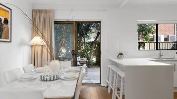 A modern kitchen with white cabinetry, a dining area with a table set for four, and a view of the outdoors through a glass door.