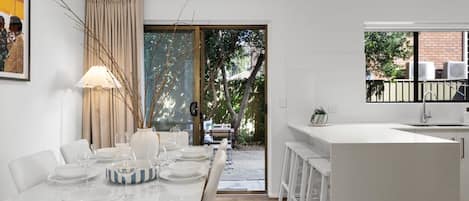 A modern kitchen with white cabinetry, a dining area with a table set for four, and a view of the outdoors through a glass door.