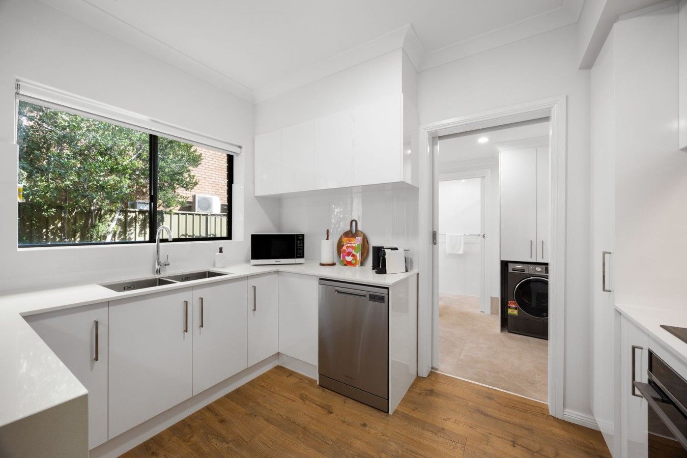 A modern kitchen with white cabinetry, a stainless steel dishwasher, and a window with a view of trees.