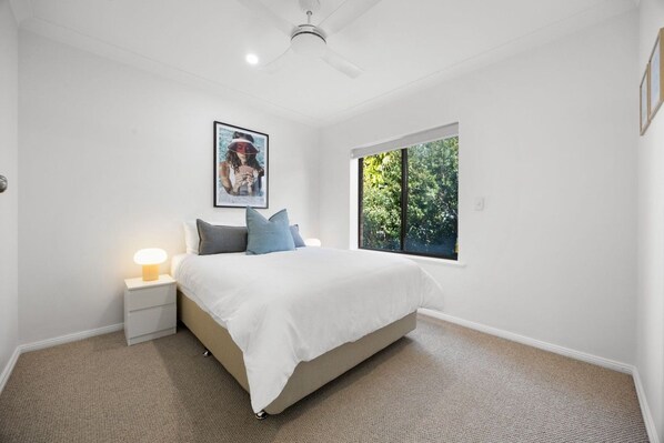 A bedroom with a bed, a nightstand, a lamp, a ceiling fan, and a window with a view of trees.