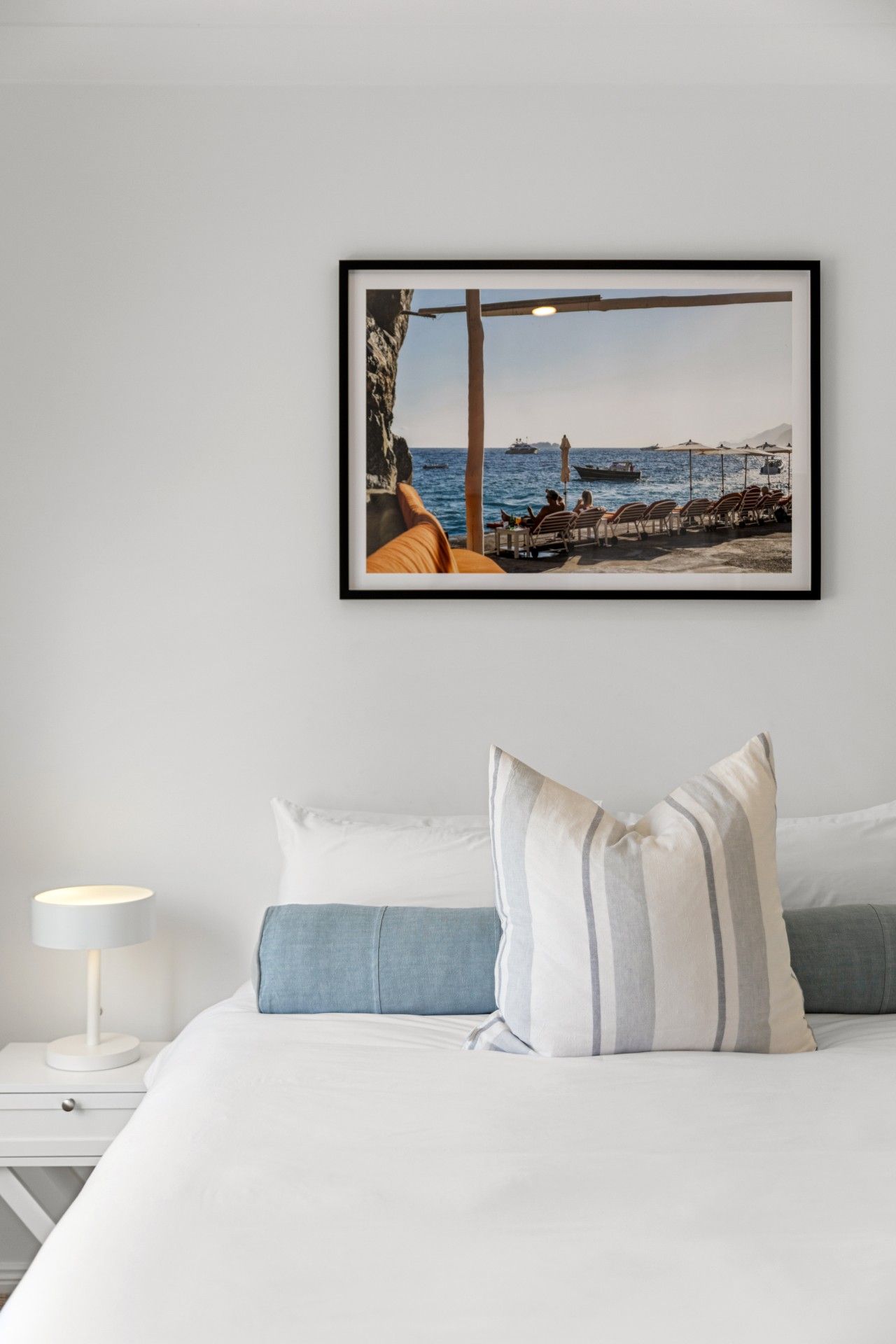 A bedroom with a bed, pillows, a nightstand, a lamp, and a framed picture of a beach scene.