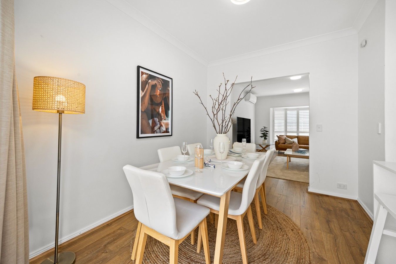 Un coin repas avec une table blanche dressée pour quatre personnes, un lampadaire et un tableau encadré au mur.