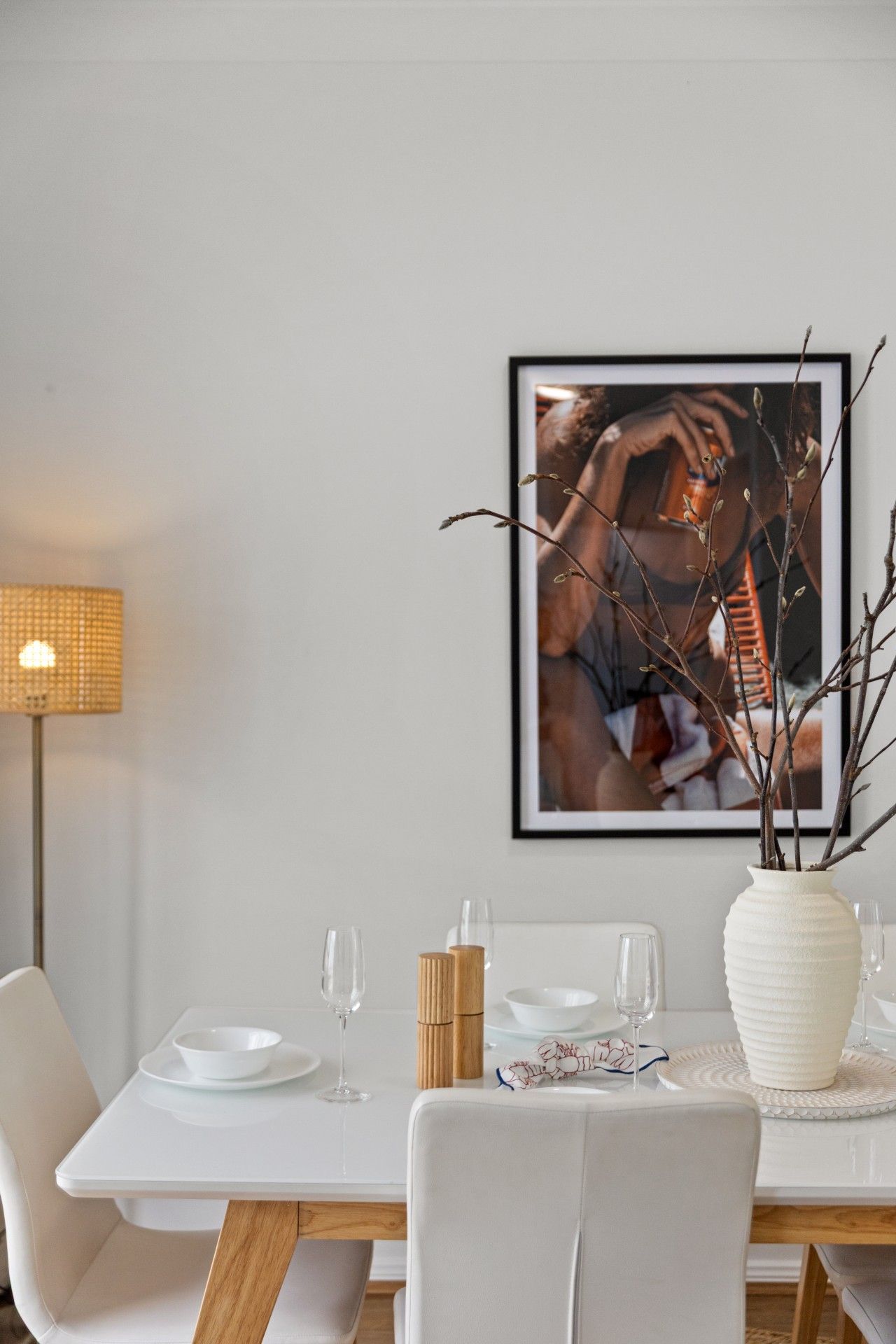 A dining area with a white table set for two, a framed photograph on the wall, and a vase with branches on the table.