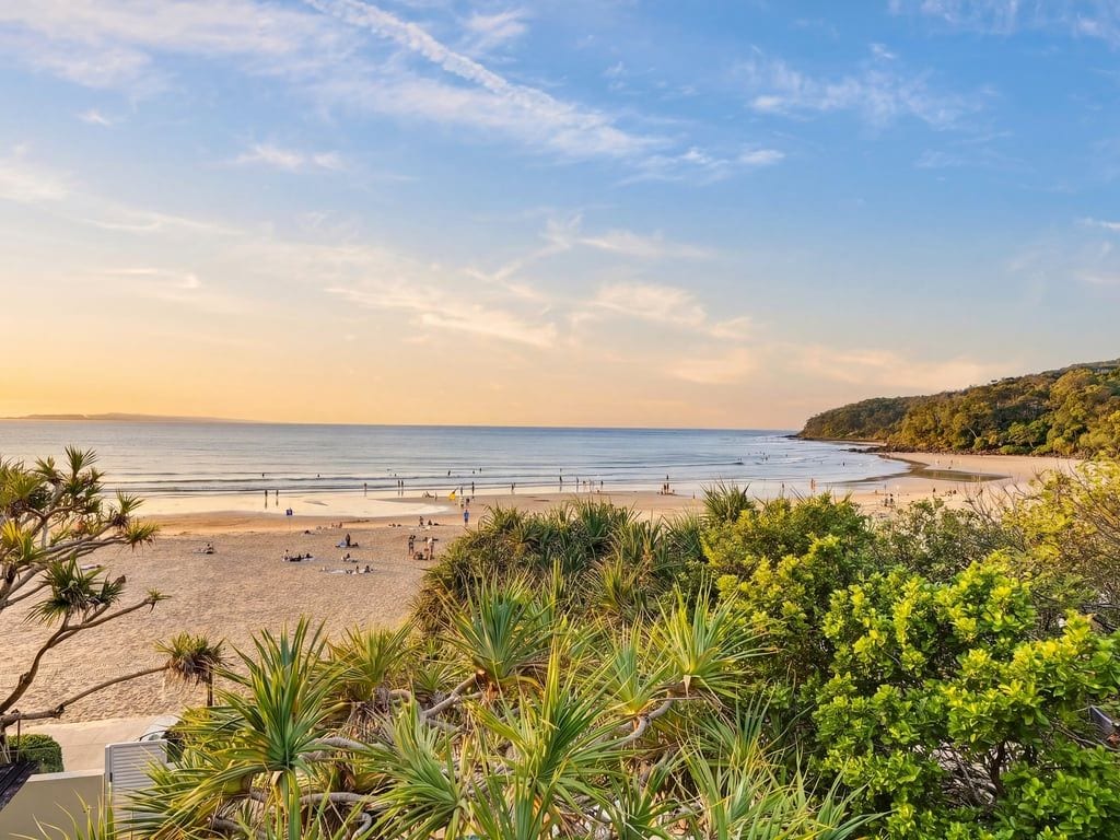 Une plage avec un ciel dégagé, un rivage sablonneux et quelques éléments de végétation au premier plan.