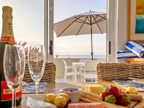Un tavolo apparecchiato con una bottiglia di champagne, bicchieri, formaggio e frutta, con vista su un balcone che si affaccia sul mare e uno ombrellone sulla spiaggia.