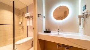 Uma casa de banho moderna com chuveiro individual em vidro, sanita branca, toucador com espelho e secador de cabelo fixado na parede.